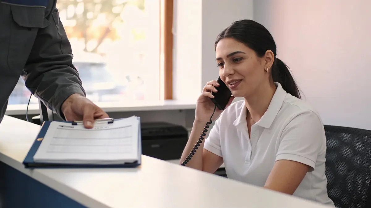 Telefonla konuşan bir kadına tezgâhın üzerinden form takılı bir klipsli dosya uzatan bir el
